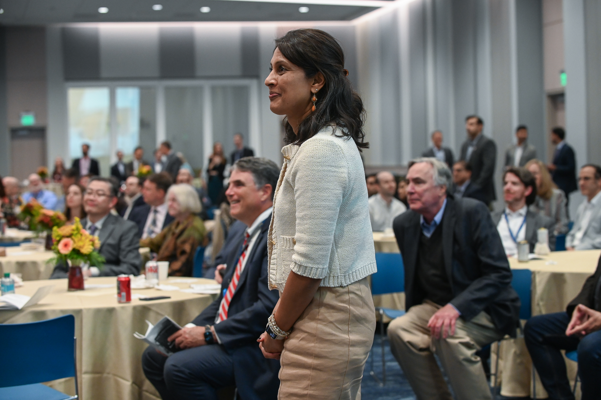 Room full of people sitting at round tables smiling with one woman standing up.