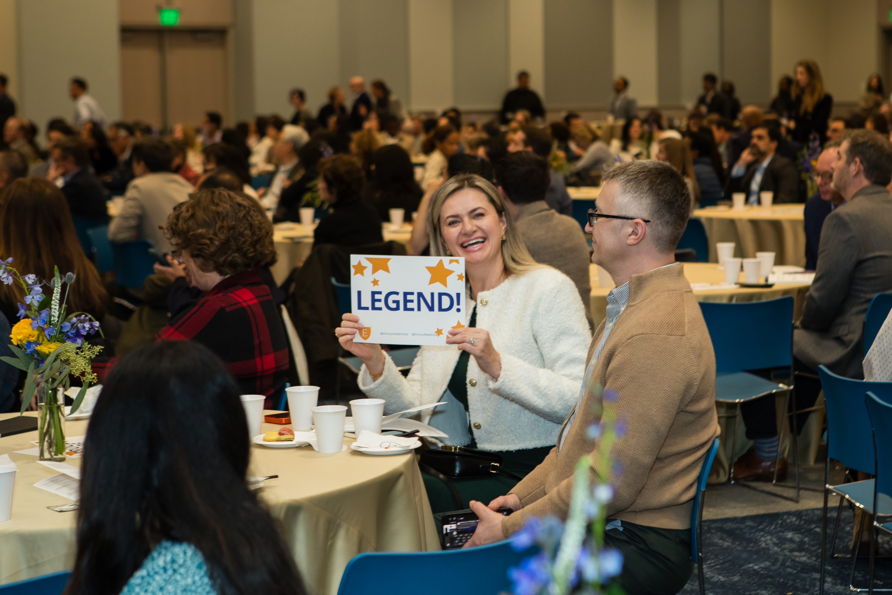 Woman smiles for a photo sitting at table holding sign that says "Legend!"