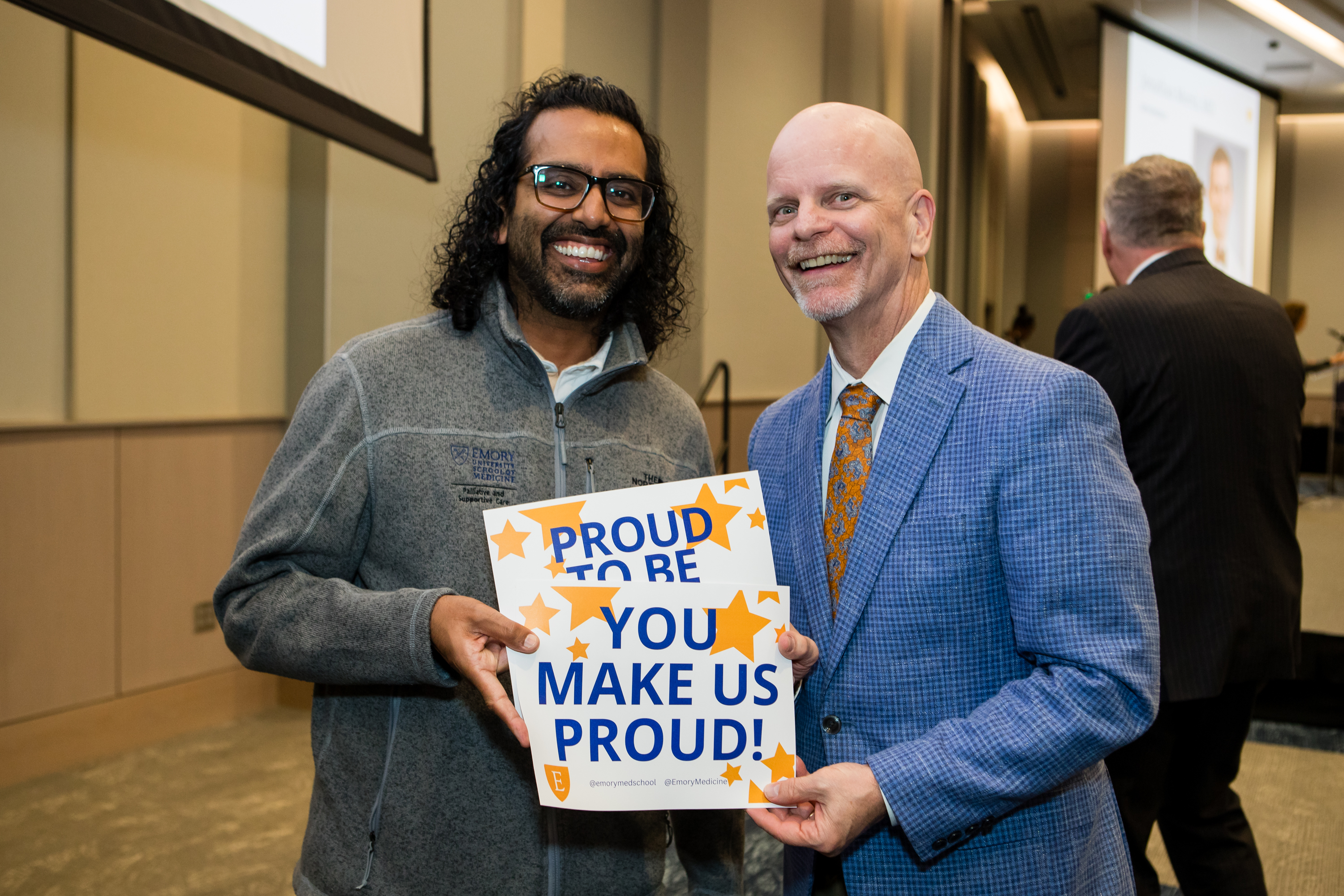 Two men smiling for a photo holding a sign that says "You make us proud!"