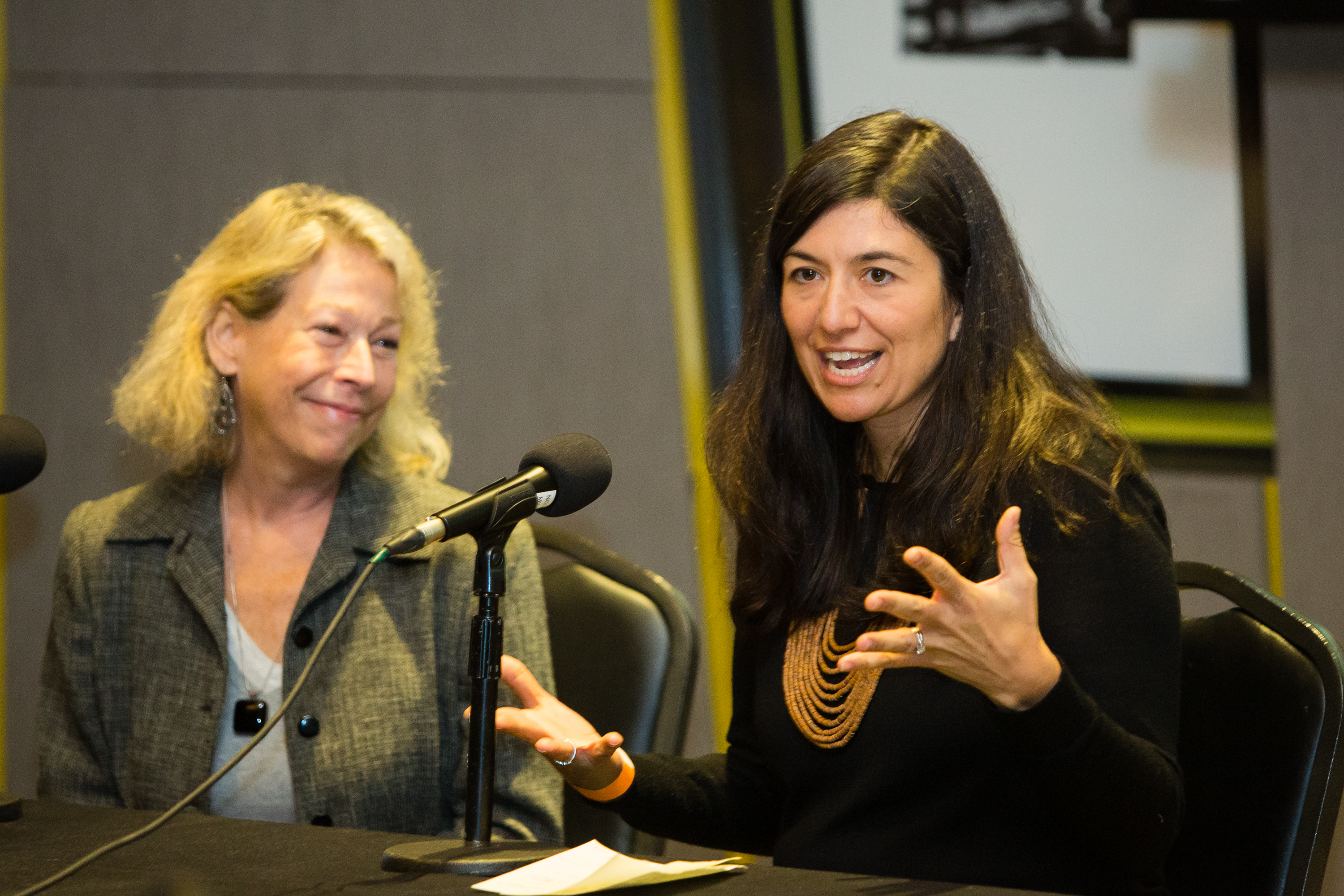 Woman speaks into a microphone while another woman smiles at her