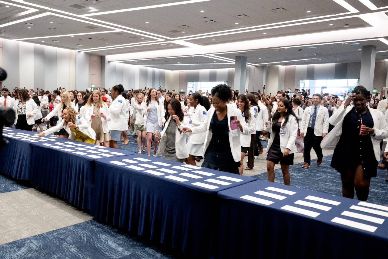 large group of people wearing white coats approaching a blue table with white envelopes