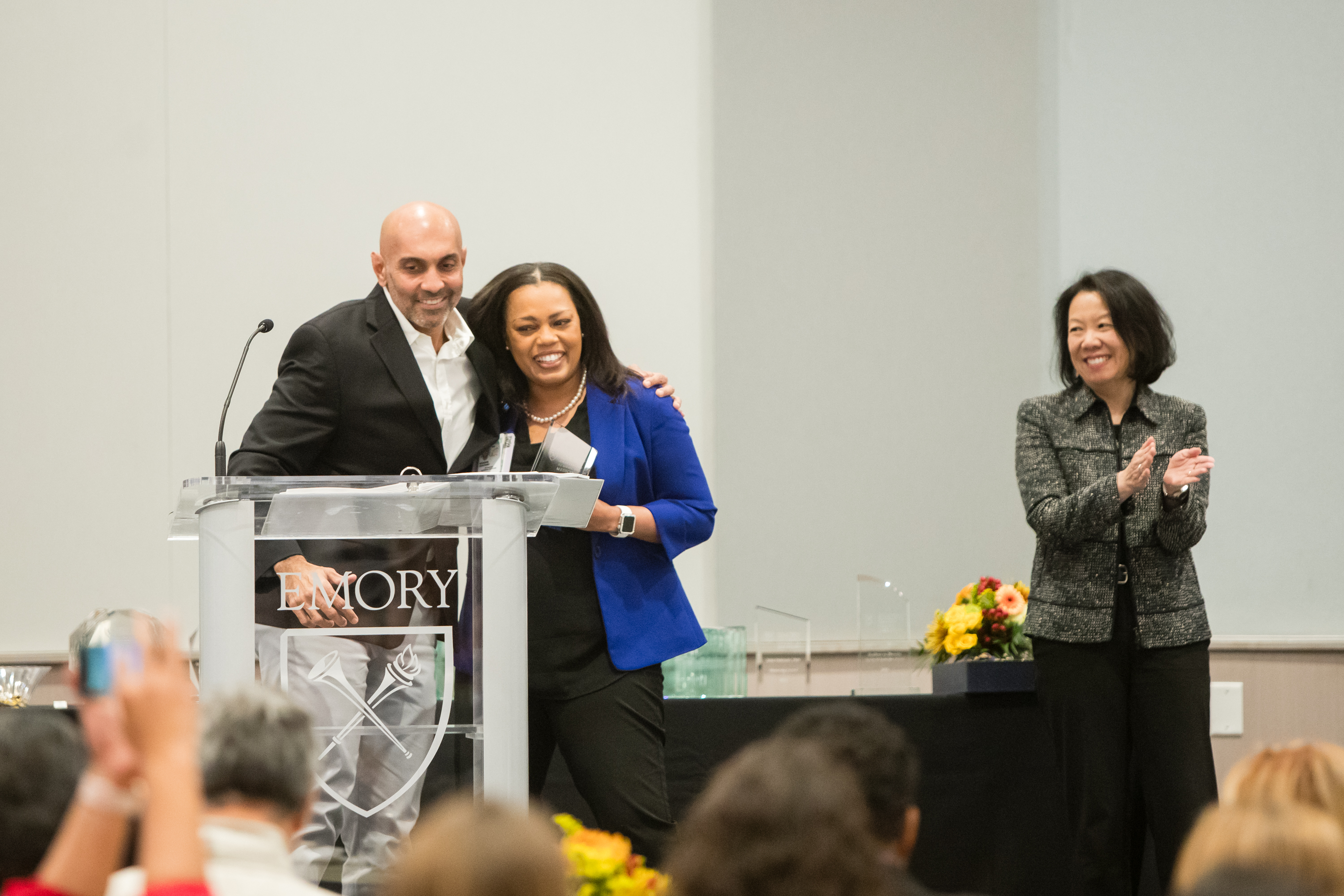 Two people smile and hug at a podium with third person off to the side clapping and smiling.