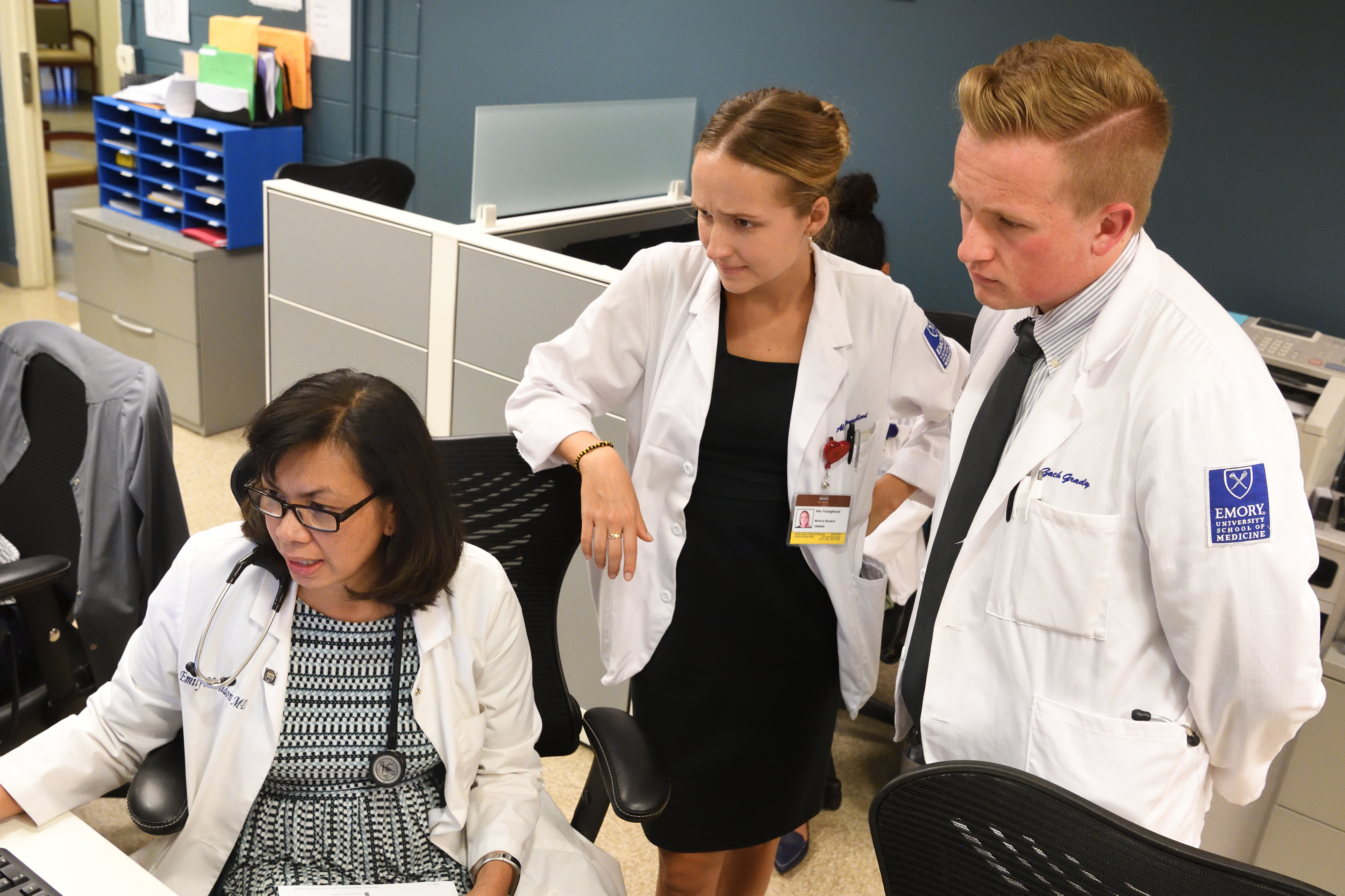 Three individuals look at a computer together in a clinical setting.