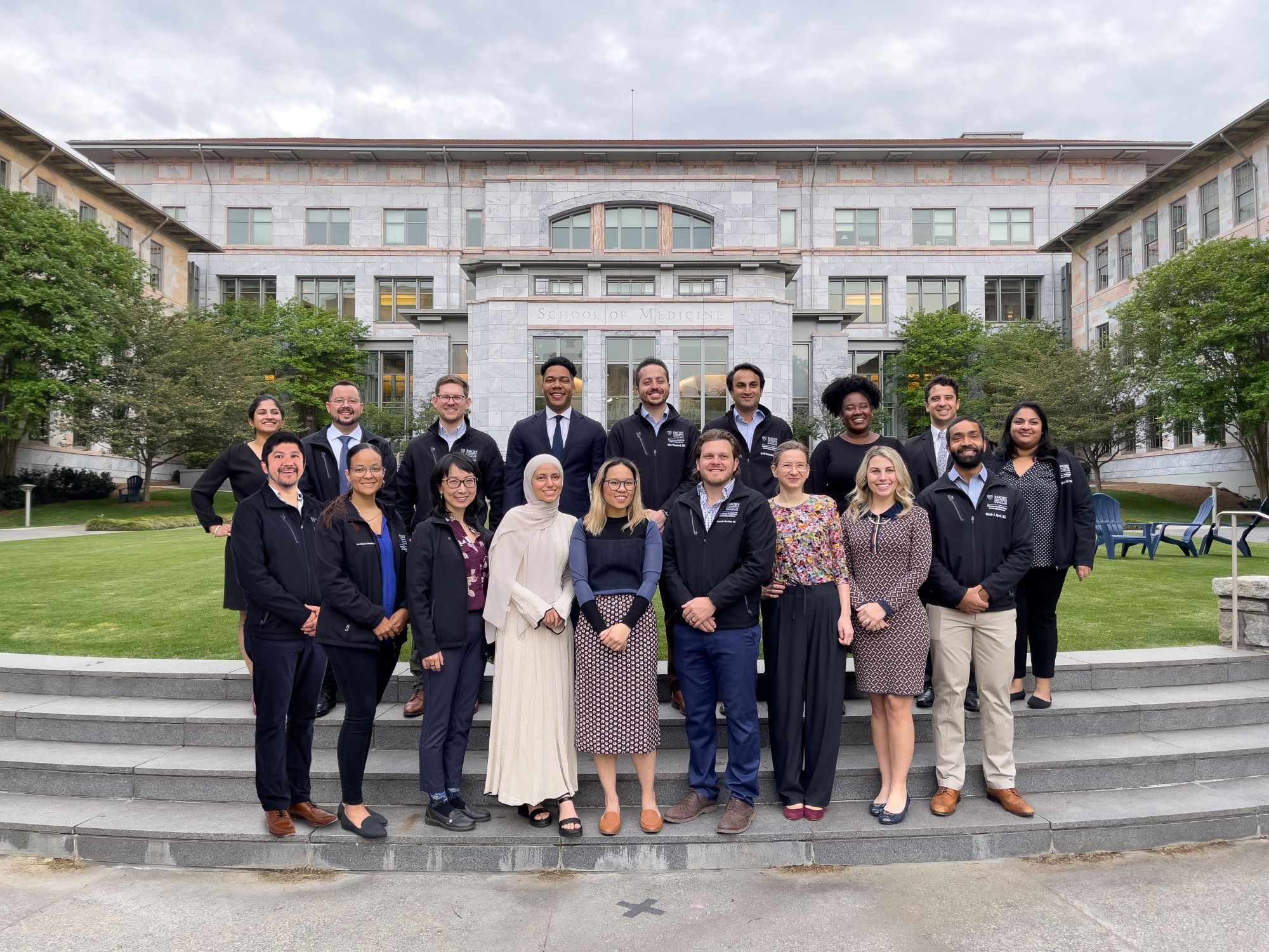 photo of two rows of people standing and smiling in front of a marble and glass building that says Emory Medicine on the front and is surrounded by small green trees and grass