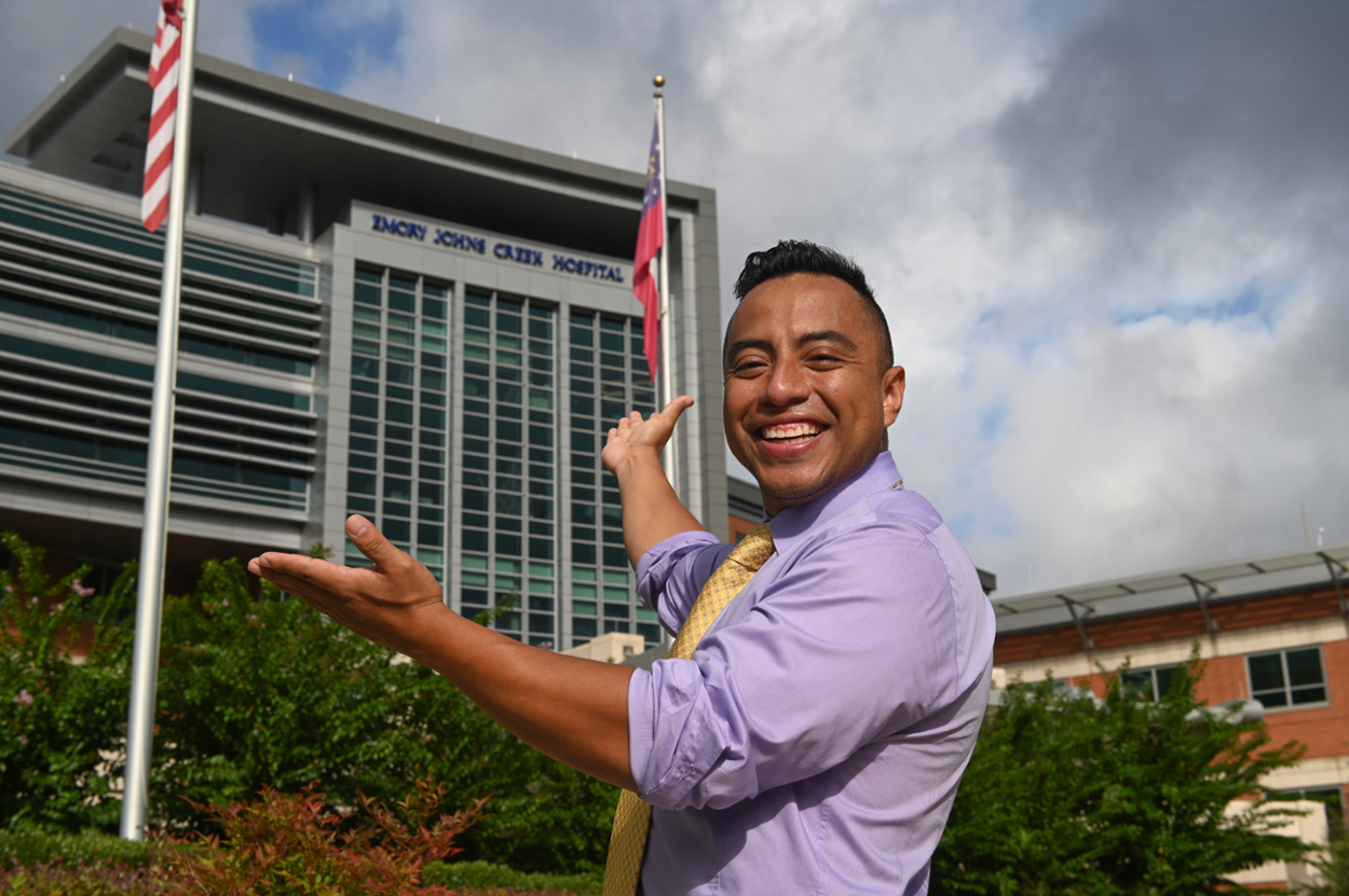 smiling man with short black hair wih his arms outstretched toward large hospital building behind him