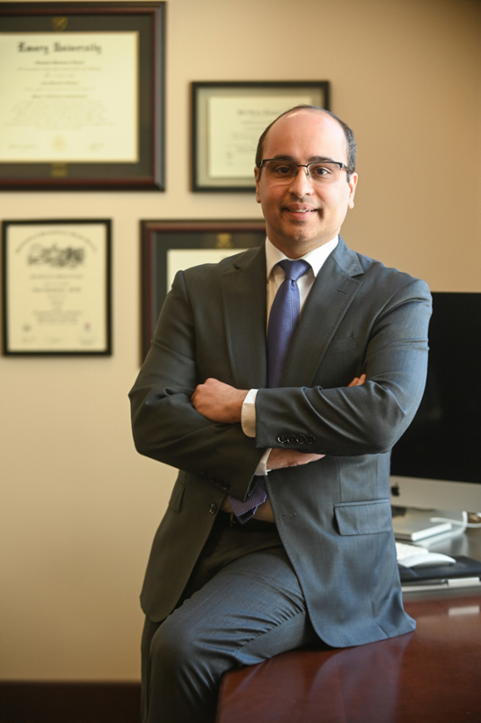 smiling man wearing glasses and dark short hair perched on edge of dark wooden executive desk with diplomas in the background