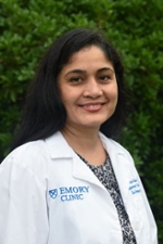 smiling woman with long black hair wearing a white doctor coat over a black and white printed top is standing in front of a green shrub background