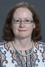 white woman wearing glasses with reddish shoulder-length wavy hair and wearing a pearl necklace and checked shirt