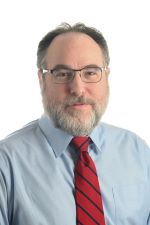 smiling person wearing spectacles and a light blue shirt with a red and black striped tie