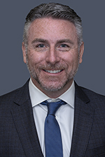 smiling man with dark and silver hair and moustache wearing dark suit coat white shirt and blue tie