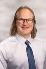 smiling man with shoulder length blonde hair and light facial hair wearing rectangular glasses and a light shirt with navy blue tie