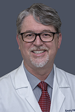 smiling man with silver and dark hair wearing dark glasses with white lab coat over blue shirt and red tie