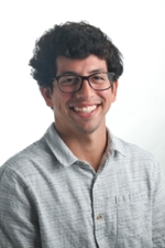 smiling person with short dark hair wearing spectacles and a grey button down shirt open at the collar