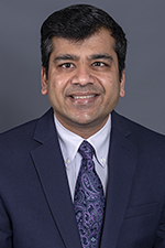 smiling man with short dark hair and wearing a white shirt, dark jacket and purple print tie