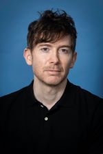 serious man with short tousled reddish brown hair and facial stubble wearing an open collared black shirt