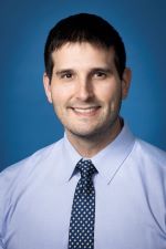 smiling person wearing a light blue shirt and blue tie with white dots