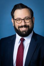 smiling man with dark short hair with beard and moustache wearing tortoise rimmed spectacles and dark suit jacket over white shirt and ruby red tie