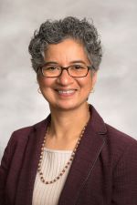 smiling woman with short curly salt and pepper hair wearing rectangular glasses and an oxblood jacket over a pale shell top layered with a gold chain