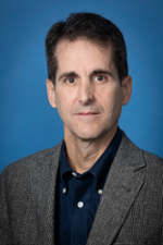 serious man with short brown hair wearing a brown tweed jacket over a black button down shirt