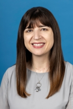 smiling woman with straight brown hair cascading over shoulders wearing a light grey top ad a silver necklace with charms dangling from a silver circle