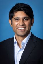 smiling man with short black hair wearing a dark suit jacket over an open collar light blue shirt