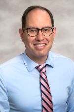 smiling man with short brown hair and wearing almost round glasses and a light blue shirt with a silver and red striped tie