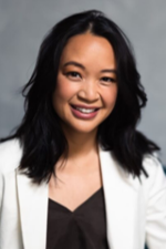 smiling person with long black hair wearing a white jacket over a brown top