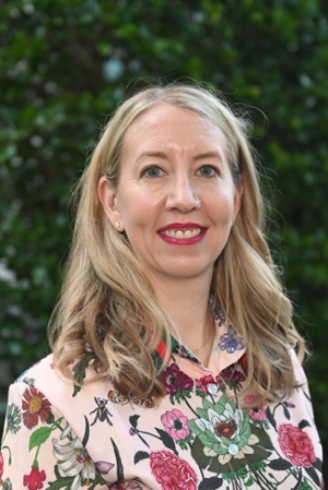 smiling woman with blond hair falling over shoulders wearing a colorful floral print top standing in front of green shrubs