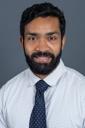 smiling person with short dark hair and facial hair wearing a white shirt with blue and grey dots and a dark blue tie with pink dots