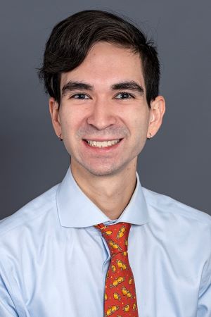 smiling person with short dark brown hair parted on the side wearing a light blue shirt and red patterned tie