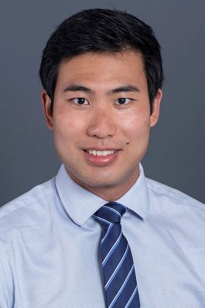 smiling person with short dark hair wearing a light blue shirt and a blue striped tie