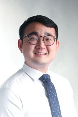 smiling man with round black-rimmed spectacles and short dark hair wearing a white shirt with a blue patterned tie