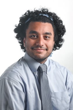 smiling man with dark curly hair held back by a light blue band and wearing a grey shirt and grey tieing 