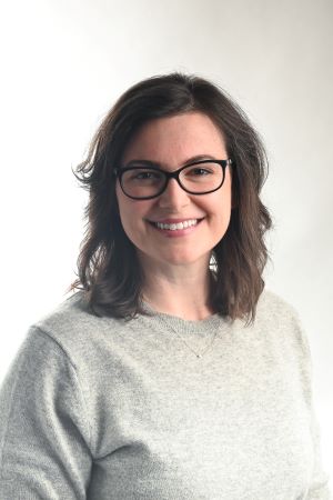 smiling woman with shoulder length wavy brown hair wearing black rimmed glasses and a light grey sweater top