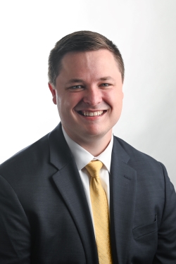 smiling man with short light brown hair and wearing a dark suit jacket over a white shirt and yellow tie