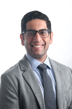 smiling man with short dark hair and dark rimmed glasses wearing a light blue shirt and gray tie under a grey suit jacket