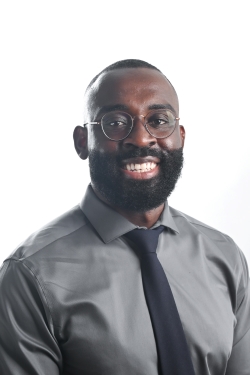 smiling man with short black hair and facial hair wearing round metal rimmed glasses and a grey shirt with a dark grey tie