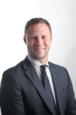 smiling man with short blond hair wearing a light shirt under a grey suit jacket and tie