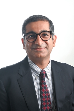 smiling man with greying dark hair and wearing black round rimmed glasses and a light checked shirt and red and black tie with a dark suit jacket