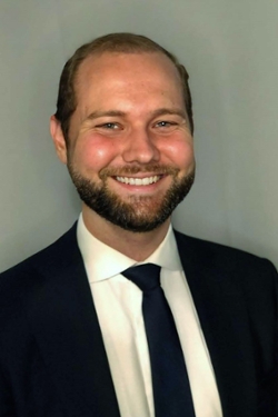 smiling person with beard and moustache and short hair wearing dark suit jacket and tie over white shirt