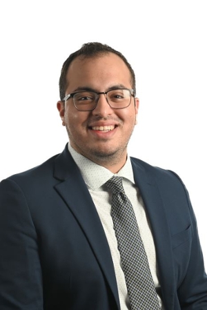 smiling person wearing spectacles and dark blue suit jacket over a cream shirt and dark patterned tie