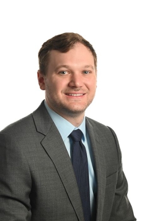 smiling person wearing a grey suit jacket over a medium blue shirt and dark blue tie