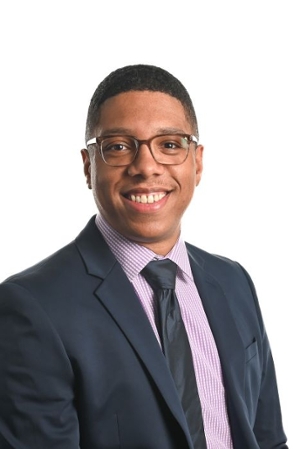 smiling person wearing spectacles and a grey suit jacket over a pink striped shirt and dark blue tie