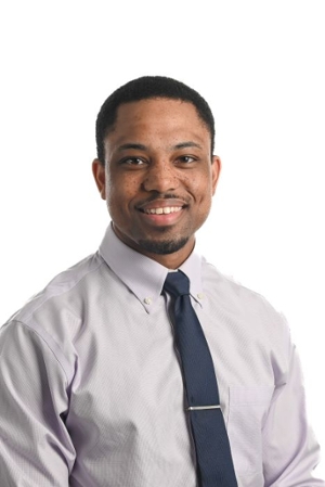 smiling person with light chin beard wearing a light grey shirt and dark blue tie with silver tie clasp