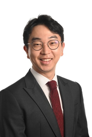 smiling person wearing round spectacles and dark grey suit jacket over a cream shirt and red tie