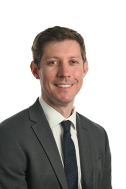 smiling person with short hair wearing a dark suit jacket and tie over a white shirt