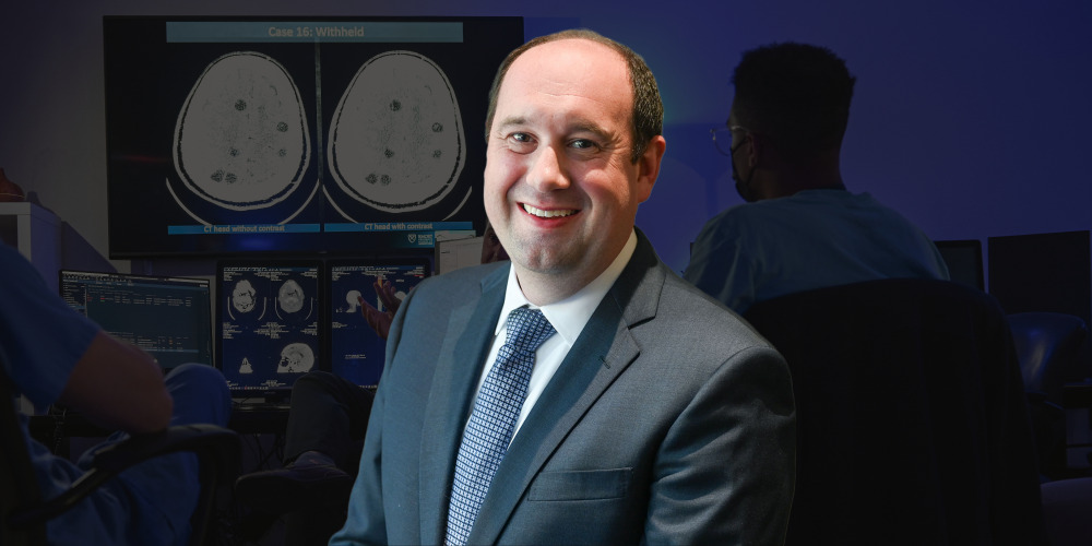 smiling person in dark suit jacket over white shirt and blue patterend tie with darkened background of people looking at radiology images