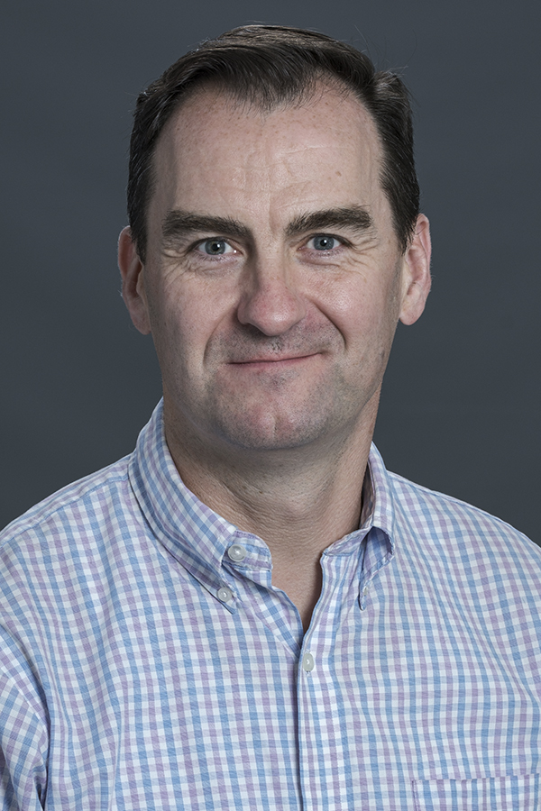 smimling man with shoart brown hair wearing a light blue pink and white checked shirt