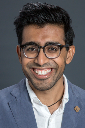 smiling man with short black hair wearing black rimmed 0spectacles and a light blue shirt under a medium blue linen jacket