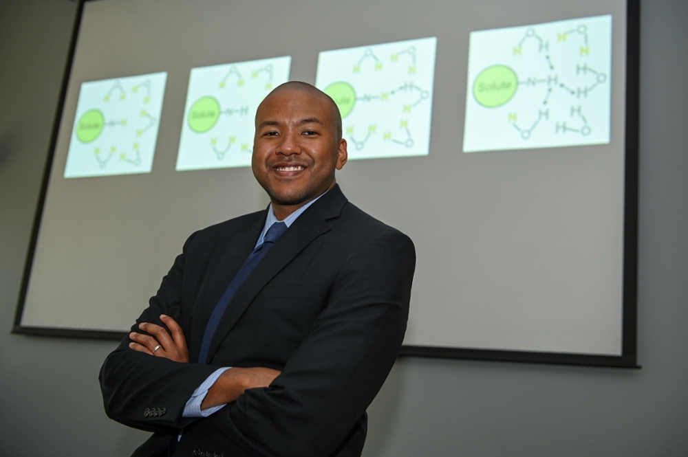 smiling man wearing dark suit with arms folded and images of a chemical equation projected behing him